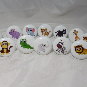 May include: A collection of white ceramic cabinet knobs, each featuring a different cartoon animal illustration. Animals include a rhino, hippo, elephant, tiger, cat, giraffe, zebra, monkey, crocodile, and lion. The knobs are round and have a glossy finish.