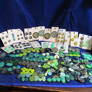 May include: A large collection of assorted buttons in various shades of green, teal, and olive. The assortment includes loose buttons of different sizes and textures, alongside several white cardboard display cards featuring sets of matching buttons.