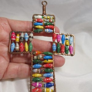 May include: A handmade cross pendant with colorful paper beads strung on copper wire. The beads are in various colors, including red, blue, yellow, and green. The cross has a loop at the top for hanging.