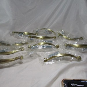 May include: A collection of gold-toned cabinet handles, each individually packaged in clear plastic. The handles have a curved design with decorative detailing. Screws are included with each handle.