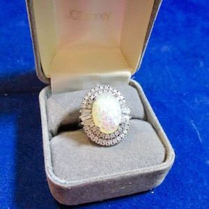 May include: An oval-shaped opal ring featuring a double halo of small round clear stones and baguette-cut stones, set in a silver-toned metal band. The ring is displayed inside a grey velvet-lined JCPenney jewelry box with a hinged lid.