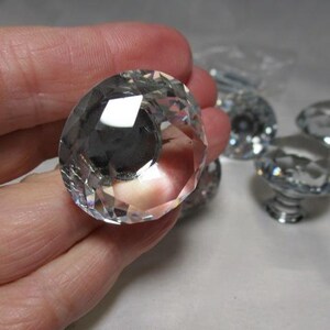 May include: Clear, faceted crystal cabinet knobs. The large, round knob is held in a hand, with several other knobs visible in the background. The knobs have a silver-colored base.