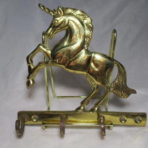 May include: A brass unicorn key holder. The unicorn is rearing up, with a long horn and flowing mane. The key holder has three hooks and a decorative design. The metal is a shiny gold color.