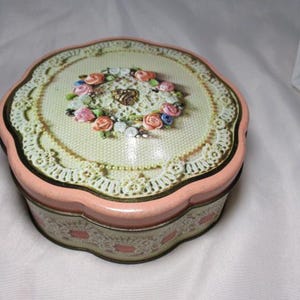 May include: A vintage, scalloped-edge tin box with a floral design. The lid features a cream-colored background with a lace-like pattern and a wreath of small, colorful flowers. The box has a pink border and base.