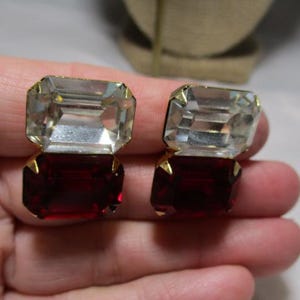 May include: A pair of vintage clip-on earrings featuring a two-tiered design. The top tier is a clear, rectangular crystal, and the bottom tier is a deep red, rectangular crystal. The crystals are set in a gold-tone metal.