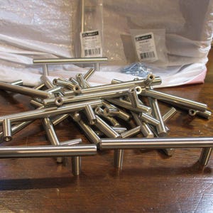 May include: A collection of brushed nickel T-bar cabinet pulls with a cylindrical design. Each handle features two vertical mounting posts with threaded holes. The hardware is made of metal with a smooth, metallic finish and a silver-toned appearance.
