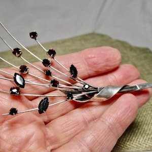 May include: A silver-toned brooch featuring a stylized floral design. The brooch has multiple thin, curved stems adorned with dark red, round and marquise-shaped gemstones. The brooch is held in a hand.