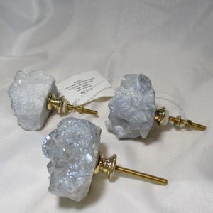 May include: Three decorative cabinet knobs with natural, light gray crystal formations. Each knob has a gold-colored base and screw, adding a touch of elegance. The knobs are designed for furniture and home decor.