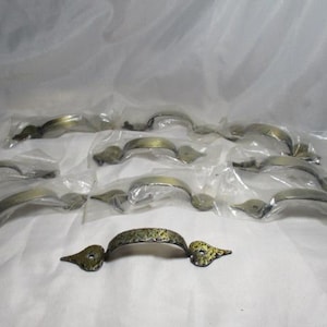 May include: A collection of antique brass drawer pulls, each in individual plastic packaging. The pulls have a curved handle design with decorative end pieces. One pull is displayed outside the packaging.