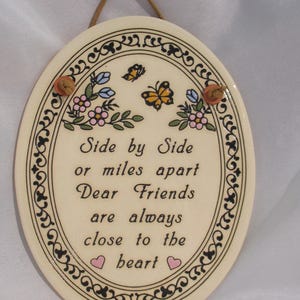 May include: Oval ceramic plaque with a cream background, featuring the text "Side by Side or miles apart Dear Friends are always close to the heart." The plaque is decorated with floral and butterfly motifs, and has a brown cord for hanging.