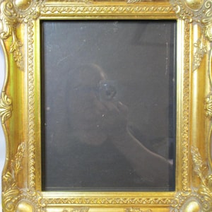 May include: A rectangular picture frame with a metallic gold finish and ornate, raised relief detailing along the edges. The frame features decorative corner accents and a smooth, inner border surrounding a dark, reflective glass center panel.