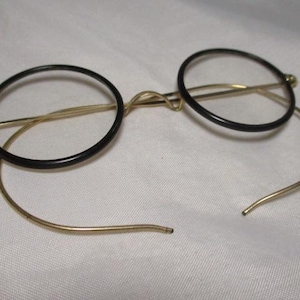 May include: Vintage round eyeglasses with black rims and gold-toned metal frames. The glasses have a classic, minimalist design, with a bridge and temples. The lenses are clear. The overall aesthetic is reminiscent of a bygone era.
