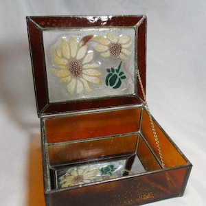 May include: A stained glass jewelry box with a hinged lid. The lid features a design of yellow and white flowers with brown centers and a green leaf. The box is brown with a gold chain attached to the lid.
