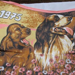 May include: A vintage textile featuring three dogs in a field of flowers. The year "1975" is printed in white at the top left. The dogs are brown and white, with pink tongues. The flowers are pink and white.