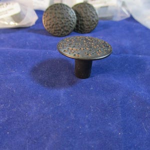 May include: A set of three round cabinet knobs with a dark matte finish and a textured, hammered surface. Each knob features a flat, circular top with a raised, irregular pattern and a cylindrical base for mounting to furniture or cabinetry doors.