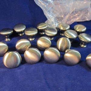 May include: A collection of round cabinet knobs with a brushed silver metallic finish. Each knob features a smooth, domed top and a tapered base. The knobs are arranged in a group, with some partially contained within a clear plastic bag.