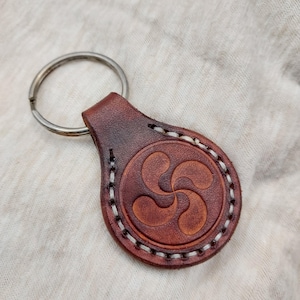 Peut inclure: Un porte-clés en cuir marron avec un anneau porte-clés argenté. Le porte-clés présente un symbole basque Lauburu en relief, cousu avec du fil blanc sur les bords. Le cuir a une riche teinte brun-rougeâtre.