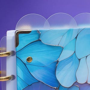 Könnte beinhalten: Nahaufnahme eines Journals mit einem Design von blauen Schmetterlingsflügeln. Das Journal hat goldfarbene Metallakzente und transparente Kunststoffteiler. Der Hintergrund ist ein Farbverlauf aus Lila und Weiß.