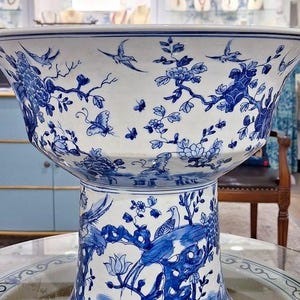 May include: A large, white porcelain bowl with a blue and white floral design. The bowl is on a pedestal with a similar design. The bowl is decorated with birds, butterflies, and flowers.