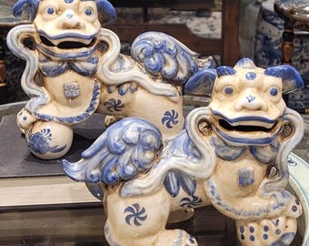 May include: Three ceramic Foo Dog statues in a cream and blue color scheme. The statues have detailed features, including stylized manes, wings, and floral patterns. The Foo Dogs are displayed on a glass surface.