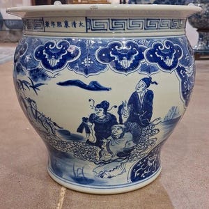 May include: A large ceramic fish bowl featuring a white base with intricate blue hand-painted illustrations of figures in a landscape. The rim displays a geometric pattern and Chinese characters. The body shows stylized clouds and foliage motifs.