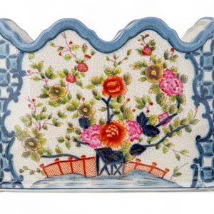 May include: An oval ceramic planter with a scalloped rim, featuring a cream-colored crackle glaze finish. The center displays a hand-painted floral motif with red, pink, and green blossoms, while the sides have a blue and white geometric pattern.