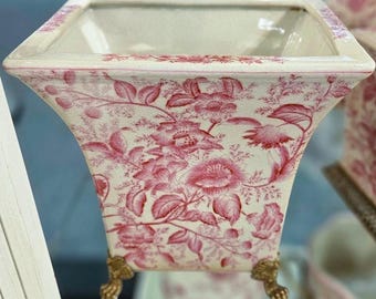May include: A square, off-white planter with a pink floral pattern. The planter has a flared shape and sits on four gold-colored claw feet. The floral design includes large blooms and foliage, creating a vintage aesthetic.