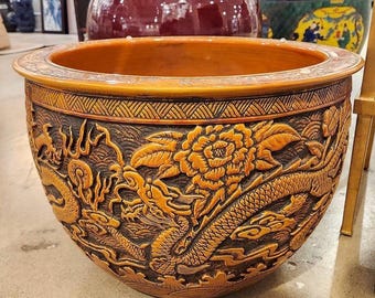 May include: A large, round, brown ceramic planter with intricate carvings of dragons and flowers. The planter has a wide, open top and a slightly flared base.