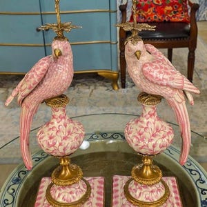 May include: Pair of pink ceramic parrot candle holders. Each features a parrot perched atop a floral-patterned vase with gold accents. The candle holders are on square bases with pink and white floral designs. The overall height is approximately 12 inches.