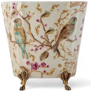 May include: A cream-colored ceramic planter with a crackle glaze finish, featuring a decorative pattern of brown birds perched on branches with pink flowers and gold accents. The vessel is supported by three ornate, gold-toned metal feet.
