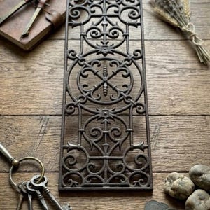 May include: A rectangular cast iron grate featuring an intricate openwork pattern of symmetrical scrolls, circles, and pointed finials. The metal surface has a dark, weathered finish with a repeating geometric design throughout the panel.