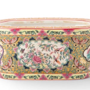 May include: Rectangular ceramic planter with a detailed floral and bird design. The planter features a yellow background with pink and blue accents, and pink handles. The central design includes birds, butterflies, and flowers.