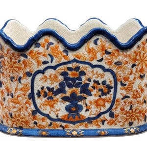May include: Oval ceramic planter with a scalloped rim, featuring a floral design in orange and blue. The planter has a blue border and a central panel with a blue floral arrangement in a vase. The background is off-white.