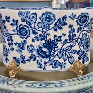 May include: Rectangular ceramic container with a blue and white floral pattern. It has gold-colored lion head handles and decorative feet. The container is placed on a round, patterned surface.