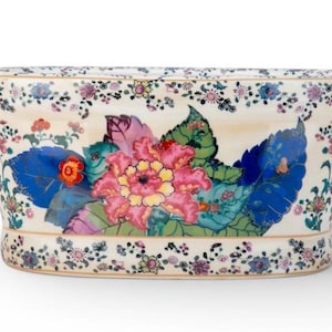 May include: Cream-colored ceramic planter with a floral design. The planter features a large pink and yellow flower surrounded by blue and green leaves. Pink handles are on each side, and the rim is decorated with small flowers.