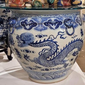 May include: Large, round, blue and white ceramic planter with a dragon design. The planter features a detailed dragon illustration, cloud motifs, and ring handles. The rim has a decorative border. A glass top is placed on top of the planter.