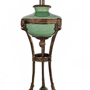 May include: A table lamp featuring a light green crackle-glaze ceramic body with bronze-toned metal accents. The lamp has a central urn-shaped section, a tripod base with ornate detailing, and a central rod topped with a brown socket fixture.