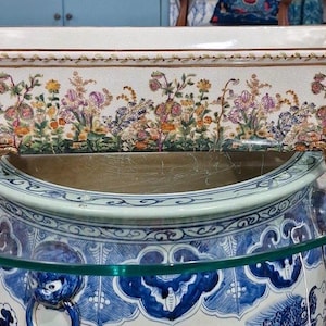 May include: Rectangular white ceramic planter with floral designs in purple, yellow, and orange. The planter has gold-colored feet and sits on a glass surface above a blue and white ceramic pot with handles.