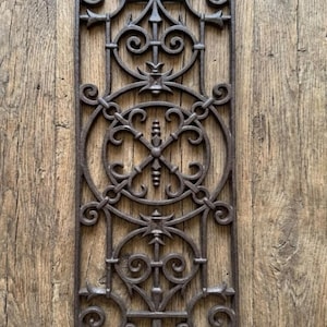 May include: A rectangular dark brown cast iron architectural grate featuring an intricate openwork pattern of interconnected circles, scrolls, and vertical finial elements arranged in a symmetrical vertical design with a textured metal surface.
