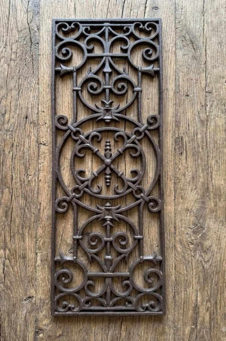 May include: A rectangular dark brown cast iron architectural grate featuring an intricate openwork pattern of interconnected circles, scrolls, and vertical finial elements arranged in a symmetrical vertical design with a textured metal surface.