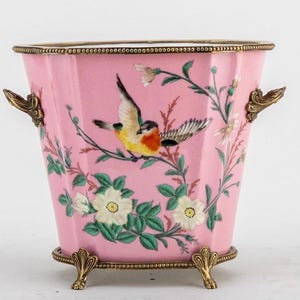 May include: A pink, octagonal decorative vase with a detailed bird and floral design. The vase has ornate gold-colored handles and feet. The bird is yellow, orange, and brown, perched on a branch with white flowers and green leaves.