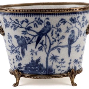 May include: A white and blue ceramic planter with a bird and tree design. The planter has a brass rim, handles, and feet. The design is reminiscent of Chinoiserie style.