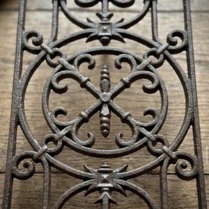 May include: A rectangular cast iron architectural grate featuring a dark brown finish. The metal panel displays a repeating pattern of circular frames, intricate scrollwork, and a central vertical spindle element with a pointed finial design.