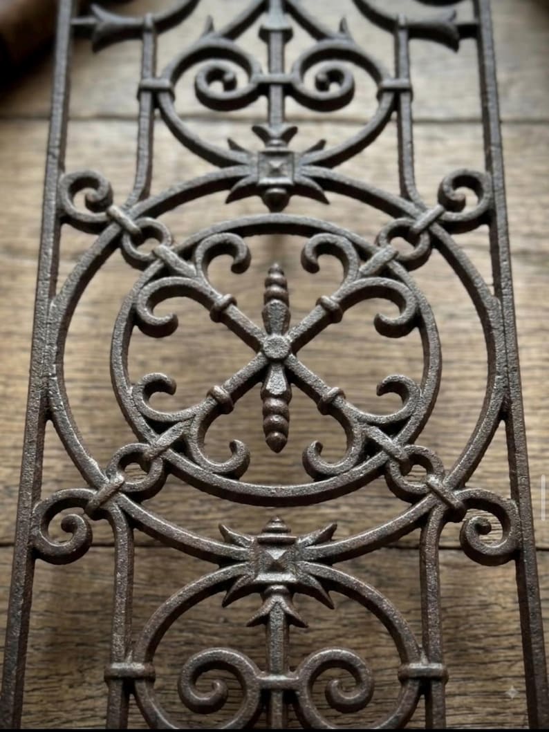 May include: A rectangular cast iron architectural grate featuring a dark brown finish. The metal panel displays a repeating pattern of circular frames, intricate scrollwork, and a central vertical spindle element with a pointed finial design.