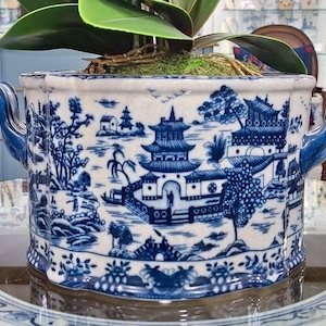 May include: A white ceramic planter with a blue chinoiserie pattern featuring pagodas, trees, and bridges. The oval-shaped vessel has a scalloped rim and two blue handles on the sides. A green plant is potted inside with visible moss.