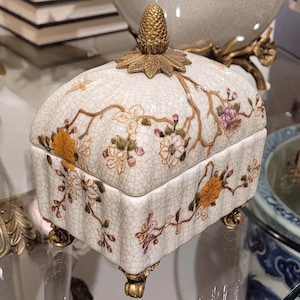 May include: A white porcelain trinket box with a gold lid and floral design. The box has four gold feet and a gold finial on the lid.