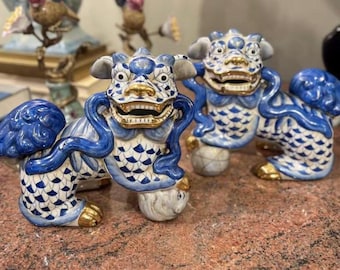 May include: Two ceramic Foo Dog statues. The statues are white and blue with gold accents. The Foo Dogs have open mouths, and are in a seated position. The statues are on a brown surface.