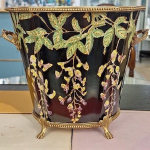 May include: A black decorative planter with gold accents and a floral design. The planter features a gold rim, handles, and feet. Green leaves and yellow and purple flowers adorn the sides of the planter.