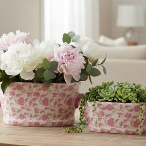 May include: Two ceramic planters with pink floral patterns. One planter holds white and pink peonies with green foliage. The other contains green succulents. Both planters have small handles and sit on a wooden surface.