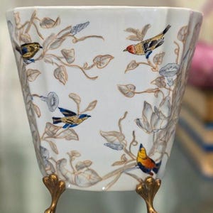 May include: A white ceramic planter with a scalloped rim and three gold-toned metal feet. The exterior features a hand-painted pattern of various small birds perched on thin, winding branches with light brown leaves and pale blue flower buds.